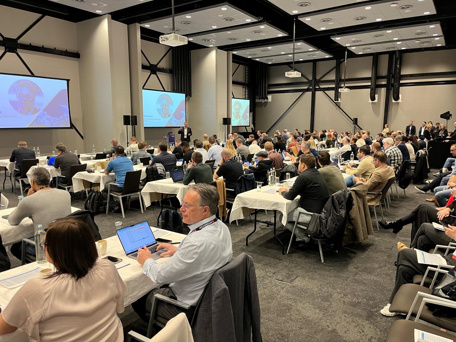 A large group of people at an event, sat on rows of tables. A speaker is stood at the front in front of a screen.