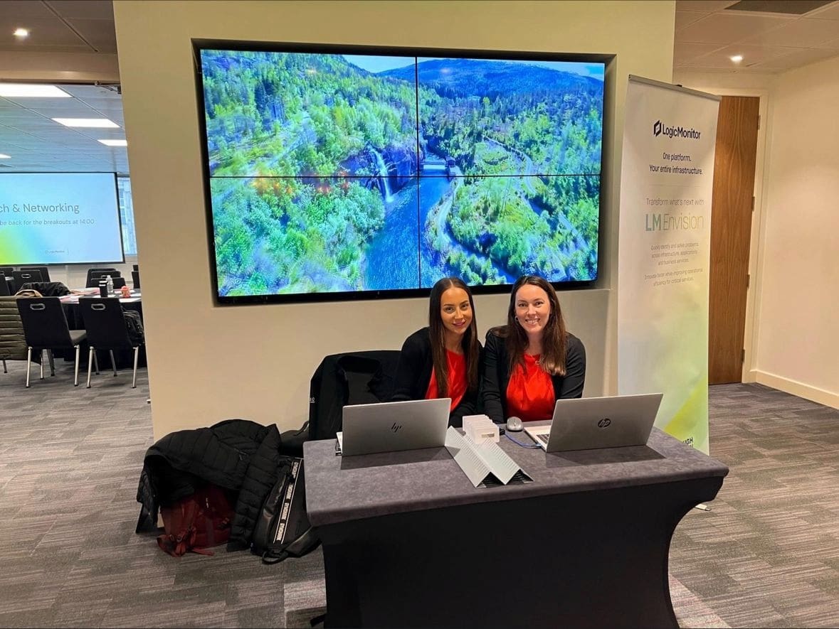 Two team members sat a desk in front of laptops, with a screen behind them
